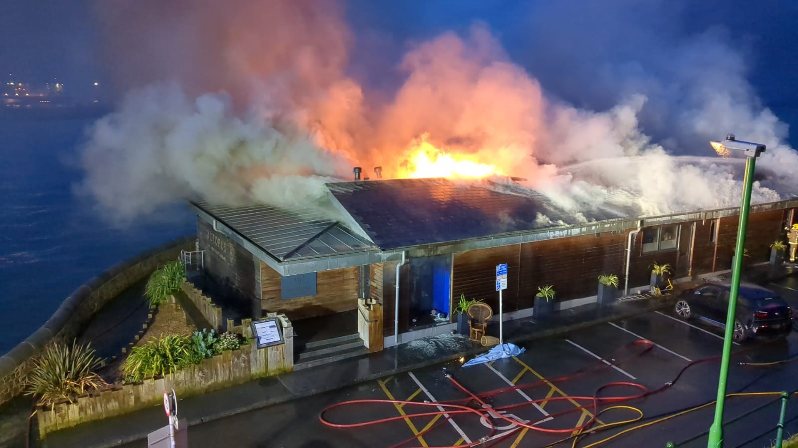 The Octopus on the night of the fire — flames and smoke pouring from the roof, fire hoses on the ground, the building lit from within by the fire, the sea black behind it